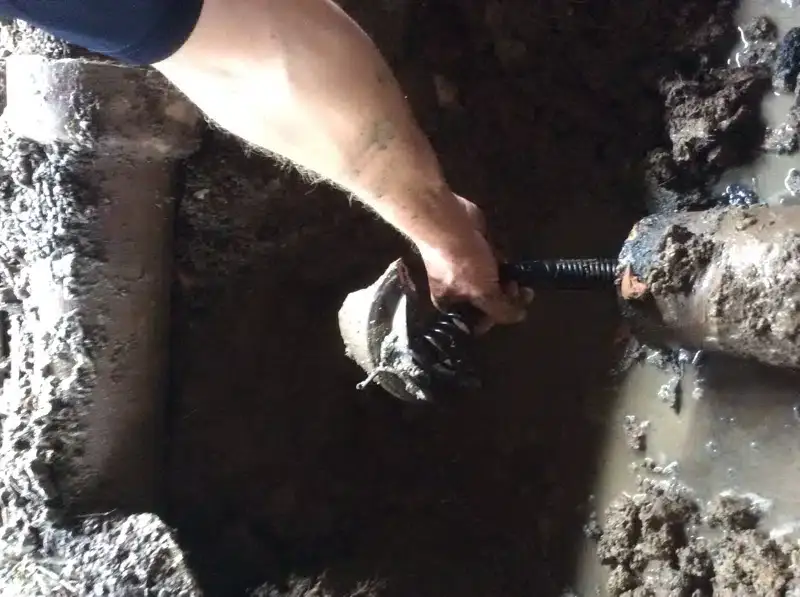Plumber clearing a broken drain pipe in a muddy excavation